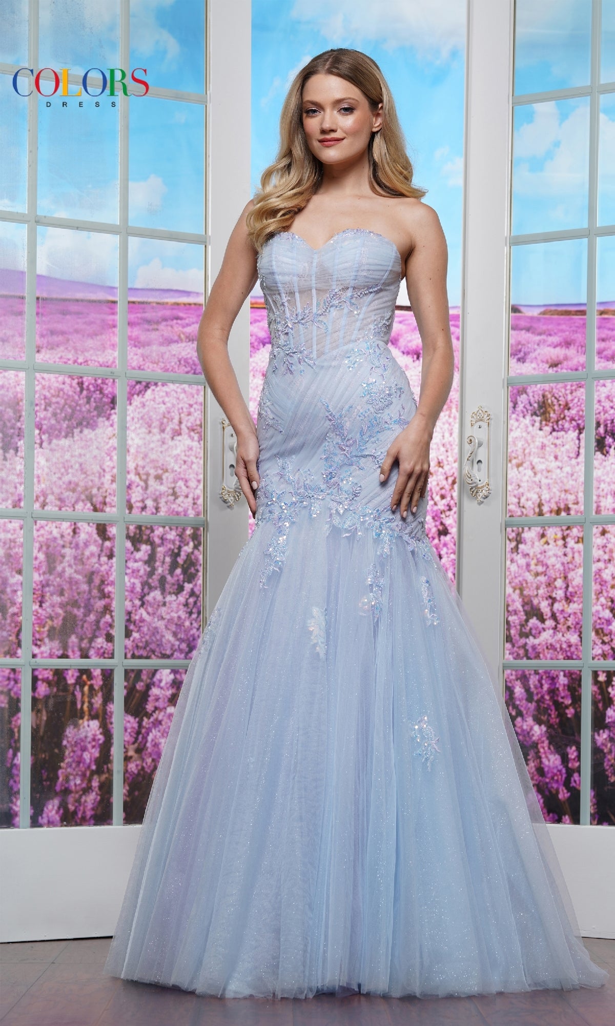 A woman wearing the Embroidered Mermaid Prom Dress: Colors Dress 3529, a light blue strapless floor-length gown, stands before glass doors with blooming lavender fields in the background.